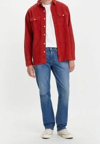 Röd kordjeskjorta med knappar och bröstfickor, bärs över en vit T-shirt och matchas med blå jeans och vita sneakers.