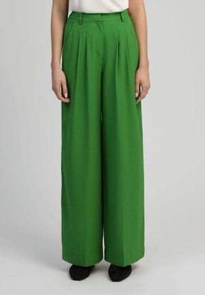 Pantalon vert taille haute à jambes larges avec plis, porté avec des chaussures noires et un haut blanc rentré, présenté de la taille aux pieds.