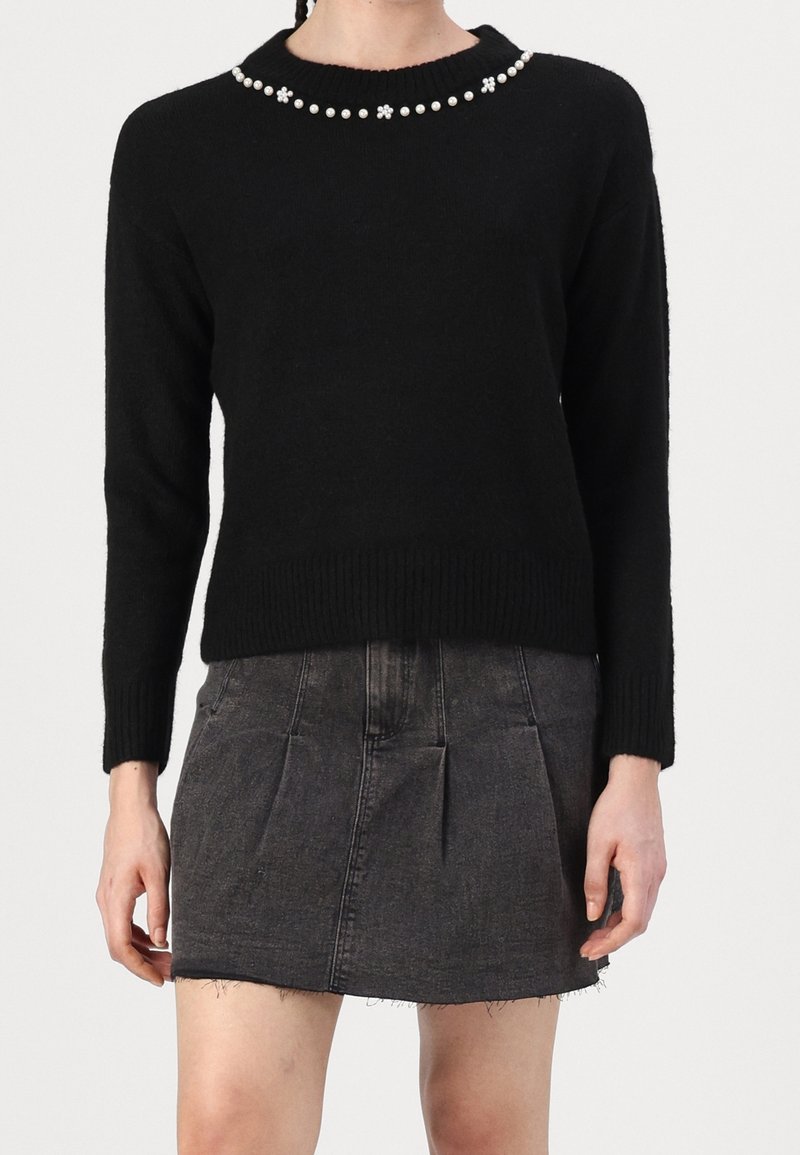 Femme portant un pull noir avec un bord en perles au niveau de l'encolure et une mini-jupe en denim gris foncé avec poches et ourlet brut.
