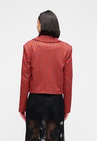 Κόκκινο cropped σακάκι με μοτίβο διαμαντιού, με κλασικό γιακά και μακριά μανίκια. Συνδυασμένο με διάφανο μαύρο φούστα από δαντέλα.