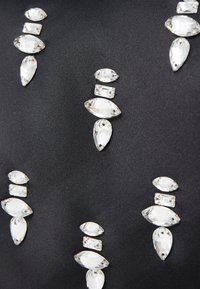 Sort svart stoff som har variasjoner av klare, fasetterte rhinestones i blad- og ovalformer, ordnet i vertikale mønstre med metalliske kanter.
