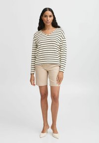 Gestreifter Pullover mit langen Ärmeln in Creme und Schwarz, kombiniert mit beigen, maßgeschneiderten Shorts. Das Model trägt spitze weiße Absätze und steht vor einem einfarbigen Hintergrund.