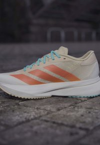 Light beige running shoe with orange stripes and blue laces on a textured pavement surface.