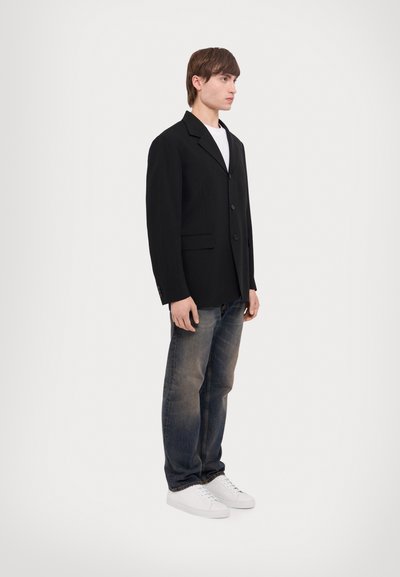 Schwarzer, taillierter Blazer mit Reverskragen, Ein-Knopfbeschluss, zwei Vordertaschen; kombiniert mit verwaschenen blauen Jeans und weißen Turnschuhen.