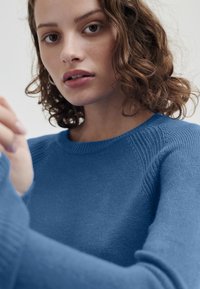 Blå stickad tröja med rund halsringning, ribbade detaljer vid axlarna och slät struktur. Närbild av modellens händer.