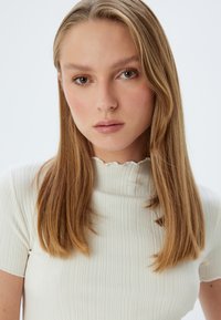 Top bianco a righe con scollatura ondulata. Il tessuto è liscio e aderente, esaltando la silhouette senza ulteriori decorazioni.