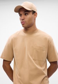 Μπεζ βαμβακερό T-shirt με στρογγυλή λαιμόκοψη, κοντά μανίκια και τσέπη στο στήθος. Συνδυασμένο με καπέλο της ίδιας απόχρωσης μπεζ με κεντημένο λογότυπο.