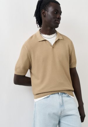 Mann med mørk hud og dreadlocks har på seg beige strukturert kortermet strikket polotrøye og lyseblå jeans, ser sidelengs med hånden bak ryggen.