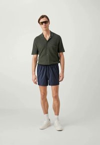 Mørk grønn kortermet skjorte med lomme, kombinert med marineblå shorts og hvite joggesko. Modellen har solbriller; omgivelsene er i nøytrale farger.