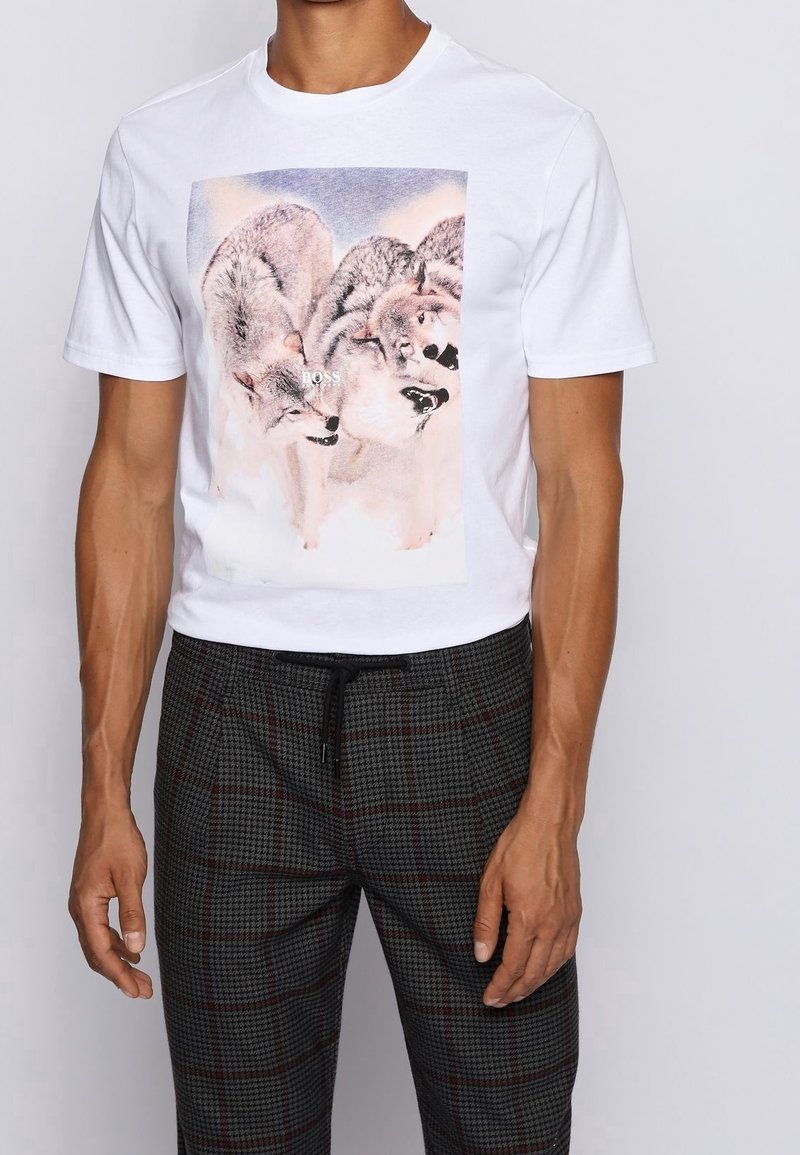 T-shirt blanc à manches courtes avec un motif de trois loups sur le devant, porté avec un pantalon à carreaux foncé avec une taille à cordon.
