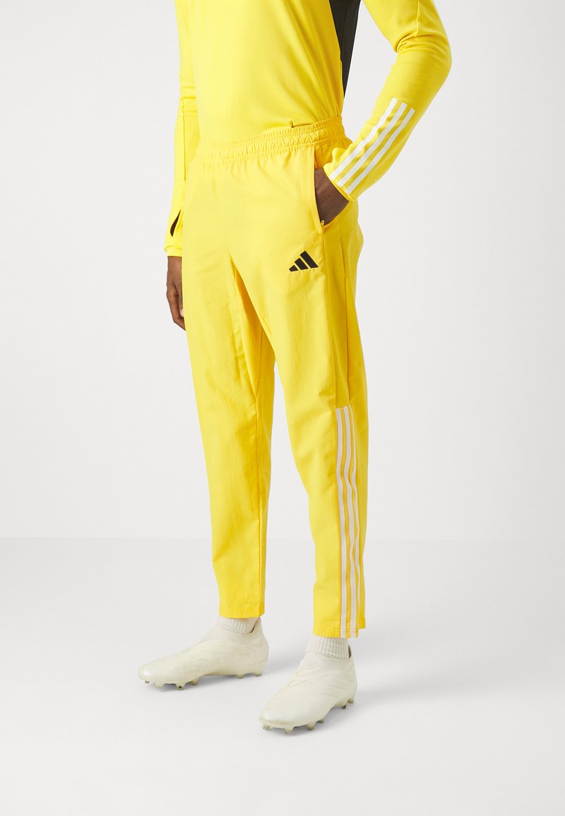 Pantalones atléticos amarillos con una cinturilla elástica, que cuentan con tres rayas blancas a cada lado y un logo de Adidas en negro. Textura suave.