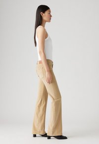 Beige utsvingte jeans laget av denim, med høy midje, kontrastsømmer og en logo-patch på baksiden. Kombinert med en hvit tanktop.