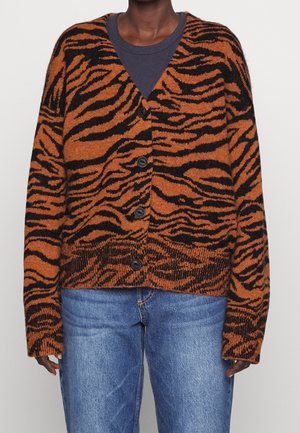 Persoon draagt een bruin-zwart geruit vest met knopen, bedrukt met tijgerstrepen, over een donker shirt en blauwe spijkerbroek.