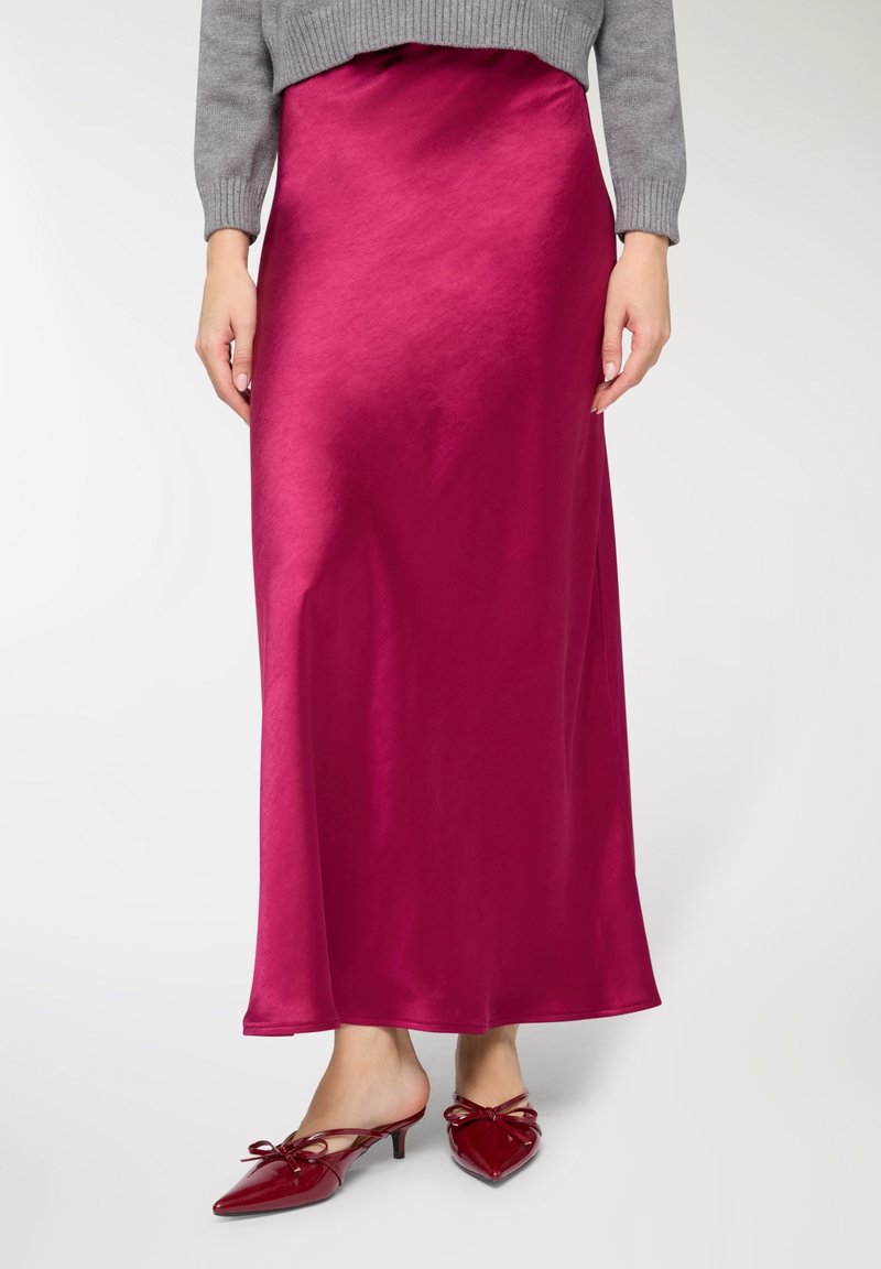 Gonna lunga in raso fucsia con una texture liscia, abbinata a mules rosse con punta appuntita e dettagli a fiocco, indossata con un maglione grigio.