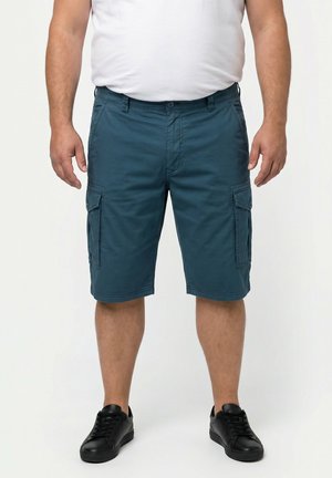 Mann trägt türkise Cargo-Shorts, weißes T-Shirt und schwarze Sneakers, steht vor einem einfarbigen hellen Hintergrund.