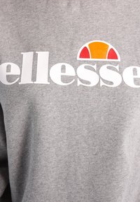 Sweat-shirt gris en tissu doux avec un grand logo blanc surmonté d'un graphique de soleil rouge et orange au-dessus du texte "ellesse".