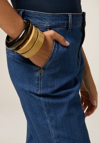 Blauwe denim jeans hebben een voorzak met een gouden metalen knoop. De hand rust binnenin de zak, versierd met gestapelde glanzende armbanden.