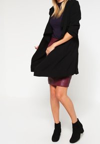 Sort trenchcoat med en afslappet pasform, båret over en mørk top og en mørkerød lædernederdel. Sorte ankelstøvler fuldender outfittet.