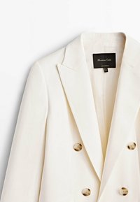 Hvid dobbeltbrystet blazer med revers, guldtonede knapper og glat stof. Etiket synlig med mærkenavn og plejeinformation.
