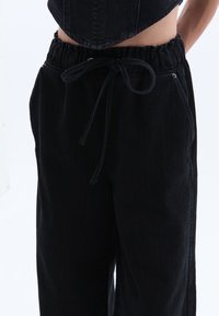 Musta farkkutakki, jossa on joustava vyötärö ja nyöri, hienovaraisella pintastruktuurilla ja sivutaskuilla.