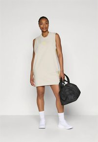 Smėlio spalvos berankovė suknelė su apvalia iškirpte, puošta geltonu logotipu ir šoninėmis plyšiais. Juoda tekstūruoto paviršiaus sportinė rankinė. Baltos kojinės ir batai.