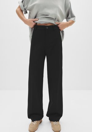 Personne portant un pantalon noir ample et des baskets beiges, soulevant un t-shirt gris pour montrer la zone de la taille sur un fond blanc uni.