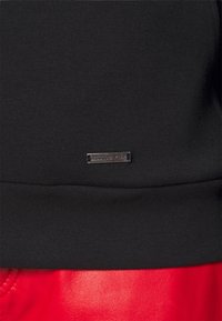 Sweat-shirt noir à texture lisse, avec une petite étiquette métallique. La partie inférieure se transforme en un matériau rouge brillant.