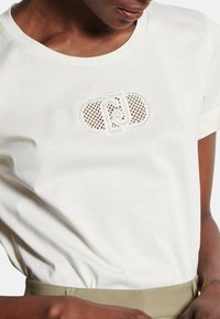 Maglietta bianca in cotone con un logo texturizzato simile a pizzo al centro. Maniche corte con orlo arrotolato e vestibilità rilassata.