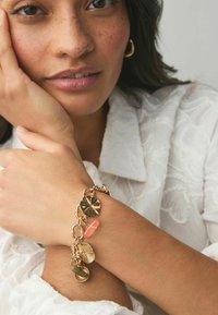 Guldtonet armbånd med forskellige charms, herunder teksturerede skiver og en lyserød accent, båret på et lys hudfarvet håndled mod en hvid baggrund.
