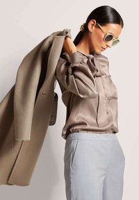 Seidene taupe Bluse mit einer Halsbinde, kombiniert mit hellgrauen, maßgeschneiderten Hosen. Das Model hält einen beigefarbenen Mantel, trägt runde Sonnenbrillen und hat das Haar glatt frisiert.