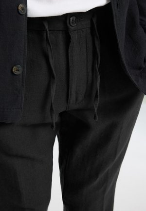Black linen pants with drawstring waist and button closure. Paired with a dark button-up shirt and white top underneath.