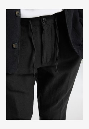 Black linen pants with drawstring waist and button closure. Paired with a dark button-up shirt and white top underneath.
