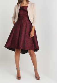 Burgundy textured dress with a flared skirt, knee-length in front, longer in back; paired with a beige cropped cardigan and silver heels.
