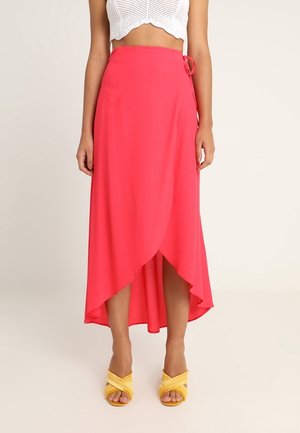 Vrouw draagt een fel koraalkleurige wikkelrok met een high-low zoom, gecombineerd met gele sandalen met gekruiste bandjes en een wit cropped gebreid topje.