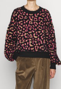 Svart, fluffig tröja med djärv rosa och tan leopardtryck; ribbade svarta manschetter och nederkant; avslappnad, överdimensionerad passform. Matchad med bruna kordflorsbyxor.