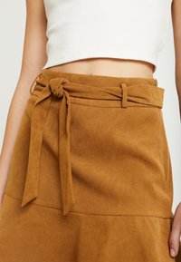 Jupe en faux suède beige avec un détail de ceinture nouée, texture lisse et design évasé. Fermeture à glissière sur le côté.