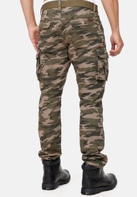 Vihreän ja beigen eri sävyissä olevat maastokuvioiset cargo-housut, joissa on useita sivutaskuja ja kapeahko istuvuus, yhdistetty mustiin saappaisiin.