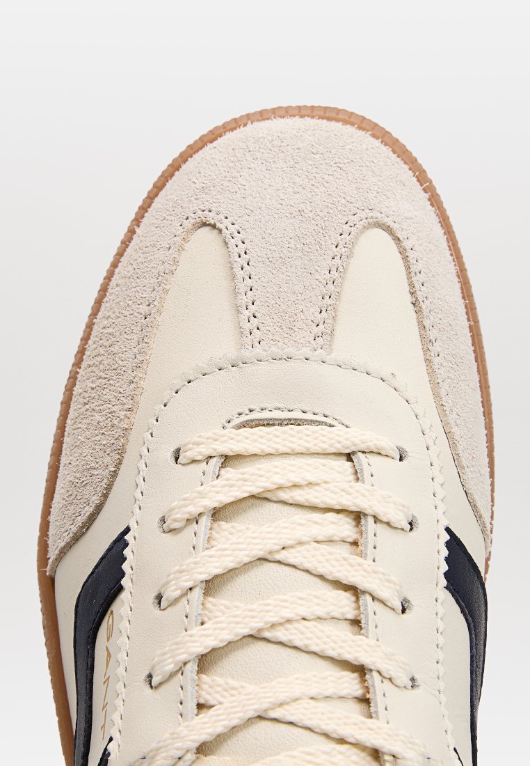 Cremefarbene und beige Sneaker mit Wildleder-Zehenschutz, weißen Schnürsenkeln und Gummisohle, die aus der oberen Vorderansicht und mit Nähdetails gezeigt werden.