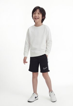 Smilende barn som står, iført hvit langermet skjorte, svarte Nike-shorts og hvite Nike-sneakers mot en hvit bakgrunn.