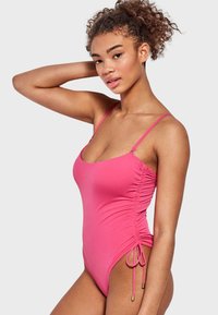Bañador de una sola pieza rosa con fruncido lateral ajustable y lazos, que presenta tirantes finos y una tela suave y elástica para mayor comodidad.