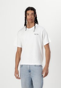 T-shirt bianco a maniche corte in cotone, con una vestibilità comoda e un piccolo logo sul lato sinistro del petto. Abbinato a jeans in denim blu chiaro.