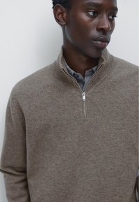 Pull en tricot texturé marron avec un col zippé au quart. Porté par-dessus une chemise grise à col, avec une fermeture éclair lisse et des détails de couture subtils.
