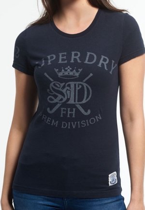 Kobieta nosząca dopasowaną czarną koszulkę z krótkim rękawem z wyblakłym sportowym logo i napisem "Superdry FH Prem Division" z przodu.