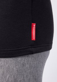 Black short-sleeve shirt with a grey, textured bottom hem and a red branded tag on the side, showcasing a simple, fitted design.