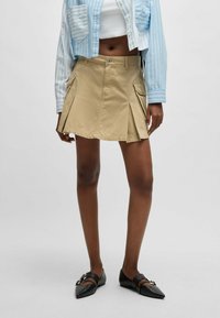 Khaki plisseret nederdel med to sidelommer, lavet af letvægtsstof. Parret med en blå stribet cropped skjorte og sorte spidse sko.