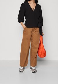 Top nero avvolgente con maniche a sbuffo, abbinato a pantaloni larghi marroni e sneakers metalliche. Tieni in mano una borsa arancione trapuntata.