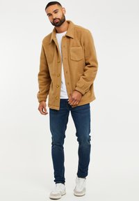 Chaqueta de forro polar color beige con cuello, cierre de botones y un bolsillo en el pecho, combinada con vaqueros azules y zapatillas blancas. Tejido limpio y texturizado.