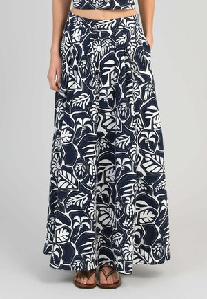 Femme portant un pantalon taille haute à jambes larges avec des imprimés abstraits de feuilles et motifs botaniques bleu marine et blanc, associé à des sandales marron.