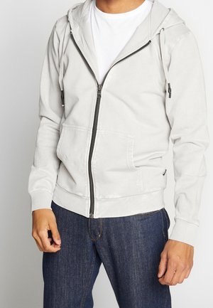 Uomo che indossa una felpa con cappuccio grigio chiaro con zip e jeans blu scuro, in piedi davanti a uno sfondo semplice, mani rilassate lungo i lati.