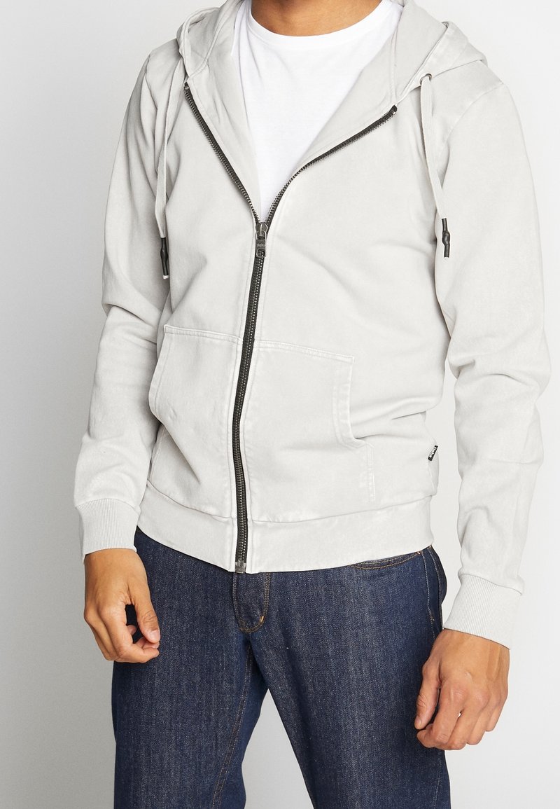 Homme portant un sweat à capuche gris clair zippé et un jean bleu foncé, debout devant un fond uni, les mains détendues le long du corps.